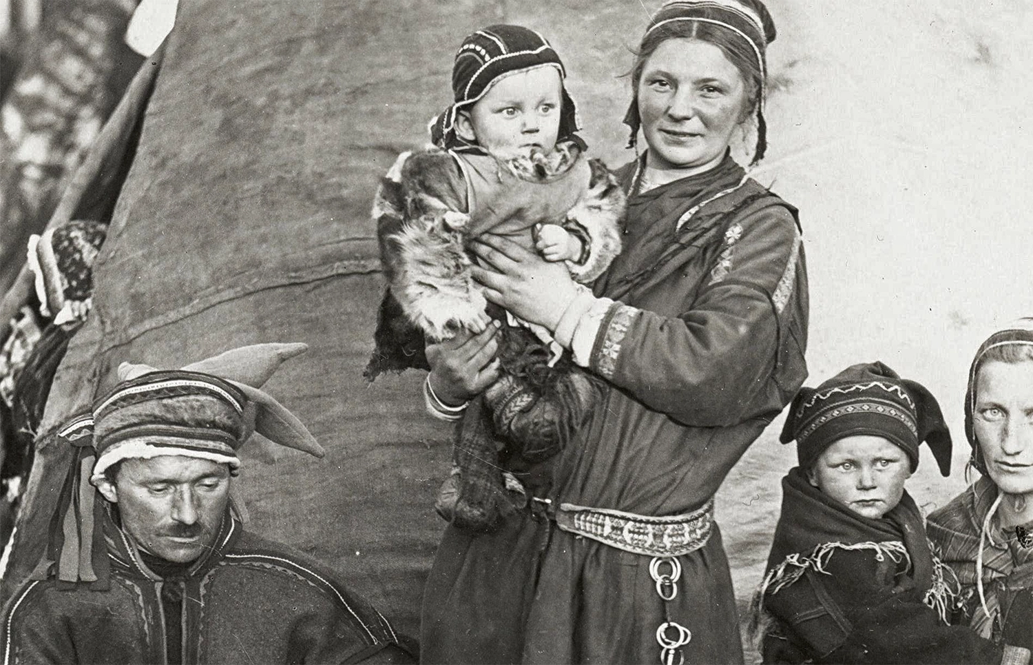 A group of traditionally clad men and women, holding small children and a dog, in front of a canvas structure.